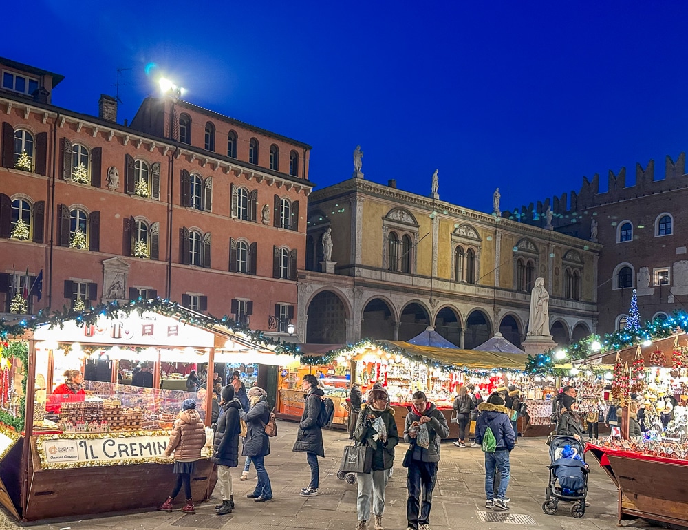 View of the Verona Christmas Market at night with wooden huts, people walking and medieval architecture in the background