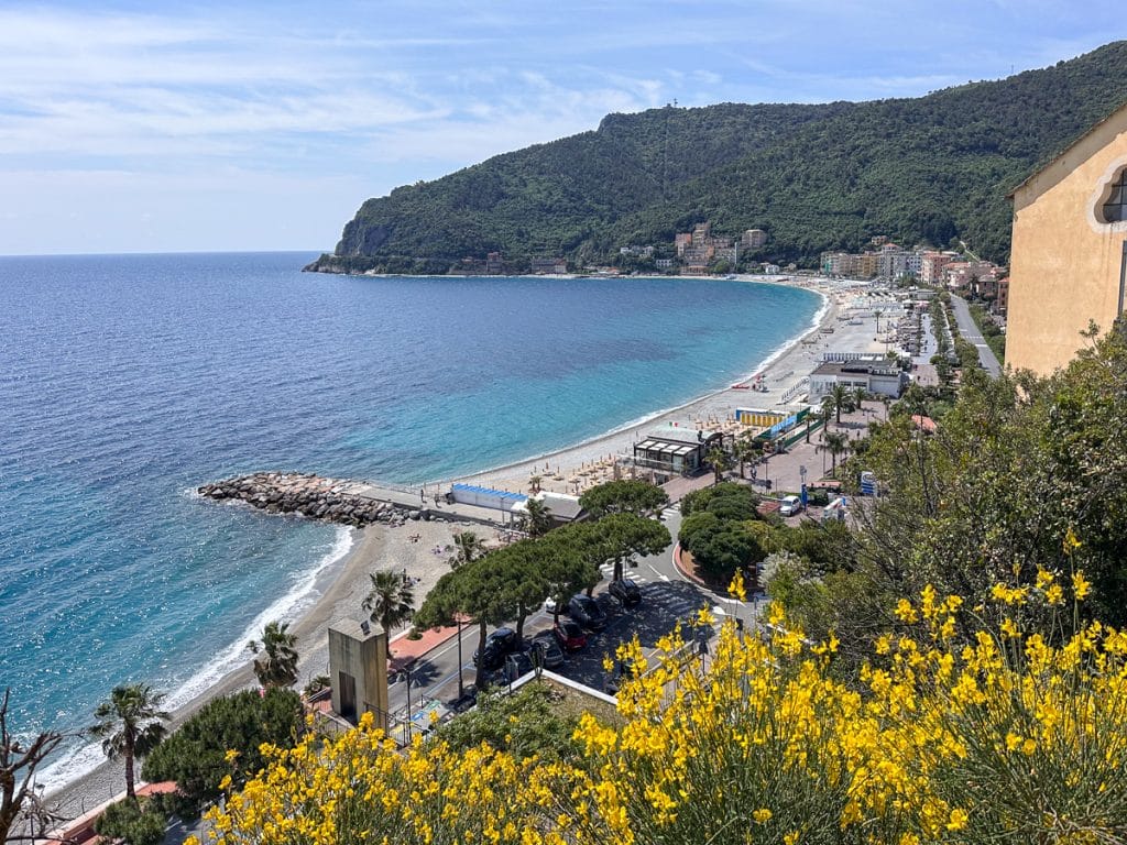 birds-eye view of Noli Italy with yellow flowers in the foreground and a turquoise sea in the background