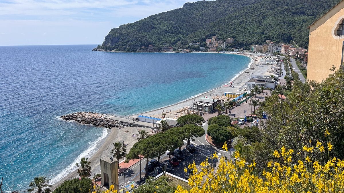 birds-eye view of Noli Italy with yellow flowers in the foreground and a turquoise sea in the background