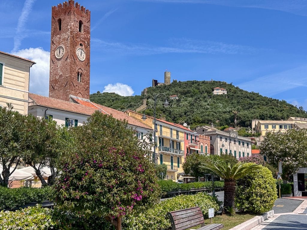 view of Noli from the beach promenade with a tall medieval tower and a castle on the hill in the background